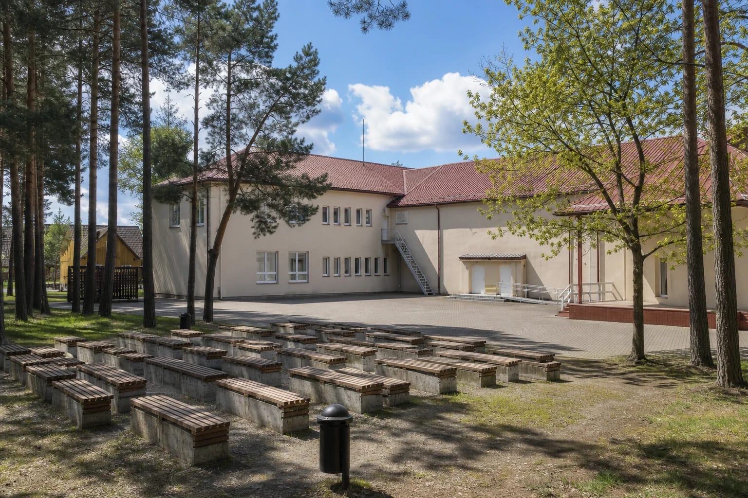 Skaudvilės kultūros centro šviesus pastatas raudonu stogu ir mediniai lauko suolai tarp pušų