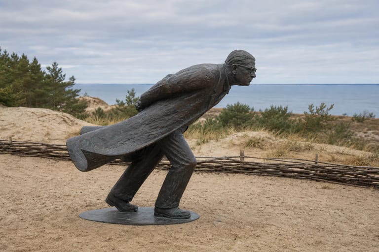 Bronzinė Jean-Paulio Sartre'o skulptūra „Prieš vėją“ palinkusi su rankomis už nugaros ant smėlėtos Parnidžio kopos, fone - Kuršių marios