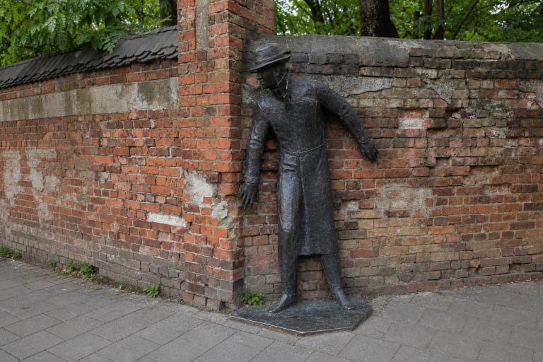 Bronzinė skulptūra „Seklys“ su ilgu paltu ir skrybėle, prigludusi prie seno raudonų plytų mūro Klaipėdoje