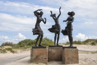 Three dark bronze Fisherman's Daughters figures on a concrete plinth among the coastal dunes of Šventoji