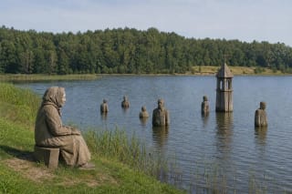 Wooden human figures and a church-tower sculpture emerging from Samylai Bay beside a green shore