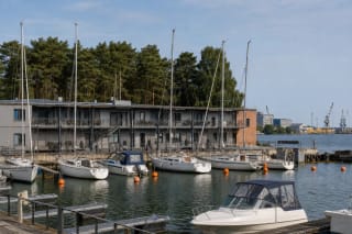 White yachts moored in a Smiltynė Yacht Club basin, with low grey buildings, an orange accent, and pine forest