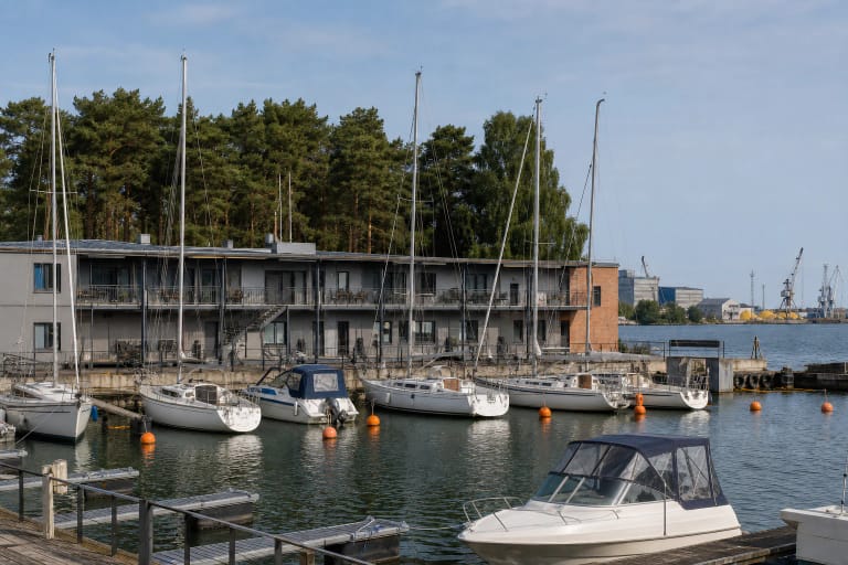 Smiltynės jachtklubo baseine prišvartuotos baltos jachtos, žemi pilki pastatai su oranžiniu akcentu ir pušynas