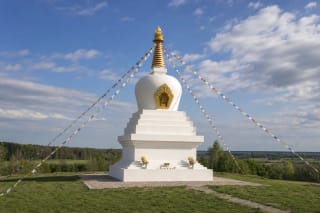 Белая буддийская ступа Ступкалниса с золотым шпилем и цветными молитвенными флажками на зелёном холме