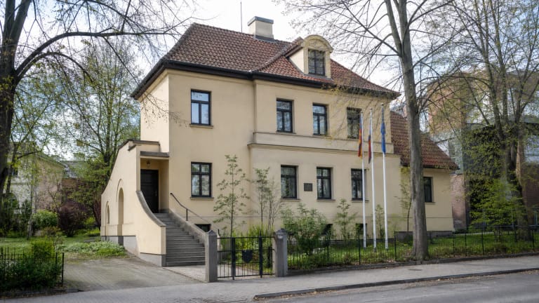 Sugiharos namų Kaune šviesus tarpukario namas su raudonų čerpių stogu, įėjimo laiptais, vėliavomis ir medžiais Žaliakalnyje
