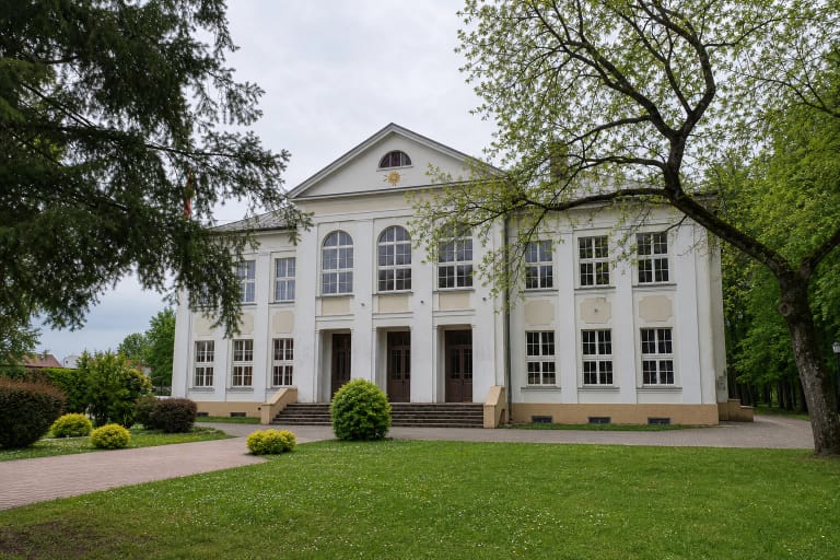 Baltas 1928 m. Švėkšnos Saulės gimnazijos pastatas su centriniu frontonu, kuriame veikia muziejus, tarp brandžių parko medžių