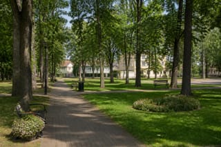 Дорожка в городском парке Швенчёниса среди взрослых деревьев, газона, клумб и скамеек