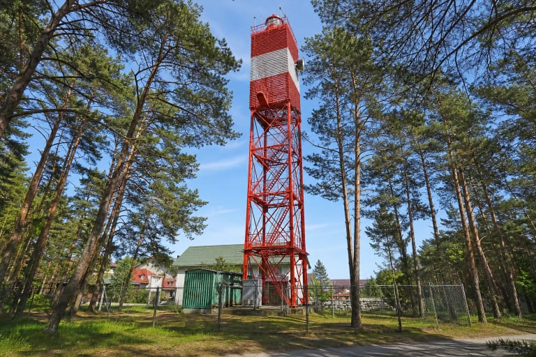 Raudonas ažūrinio metalo Šventosios švyturys su plačia balta juosta viršuje, kylantis tarp pušų už techninės teritorijos tvoros