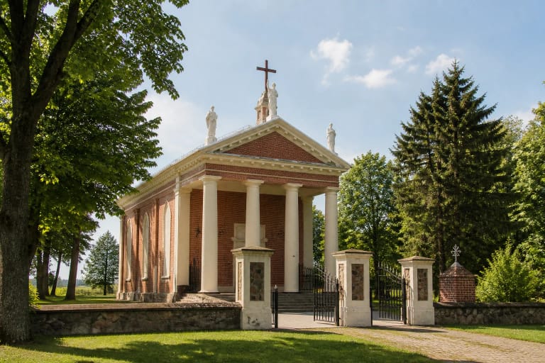 Taujėnų Šv. Kryžiaus Išaukštinimo bažnyčia su raudonų plytų sienomis, šviesiu portiku, bokštu, vartais ir medžiais