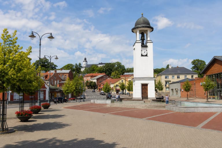 Telšių senamiesčio Turgaus aikštė su baltu laikrodžio bokštu, fontanu, raudonų ir pilkų trinkelių danga bei ant kalvos kylančia bažnyčia