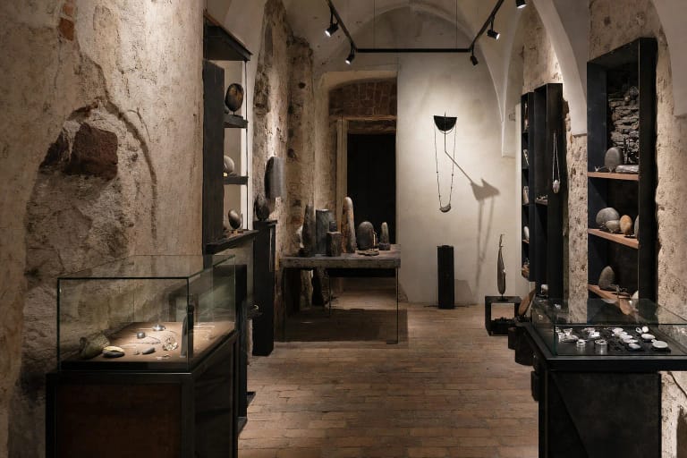 Terra Recognita galerijos skliautuotas interjeras su tamsiais akmens ir metalo objektais ant šviesių pjedestalų