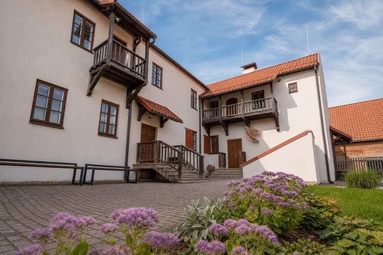 Arnetų namo kiemas su baltais tinkuotais korpusais, čerpių stogais, medinėmis galerijomis ir žydinčiais šilokais