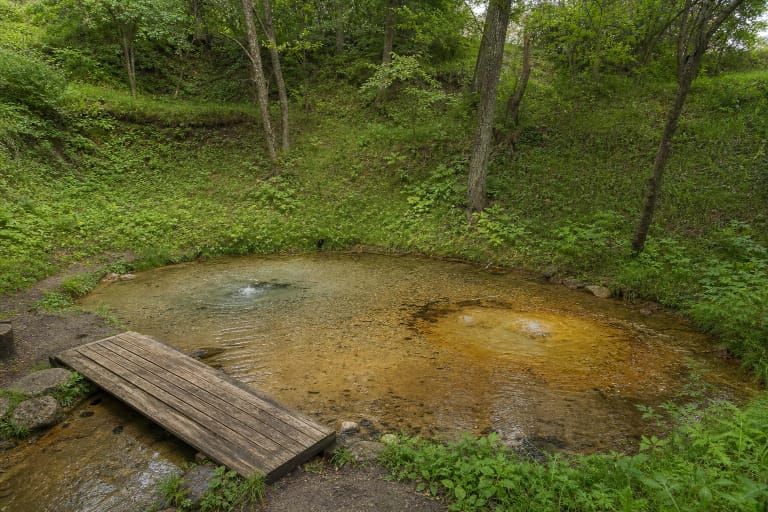 Shallow sandy basin at Šmitos Spring with a pale spring eye, a yellow-brown spring eye, and a timber footbridge