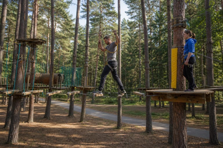 Druskininkų UNO Park lankytojas su saugos apraišais eina kabančiomis medinėmis lentomis tarp pušų