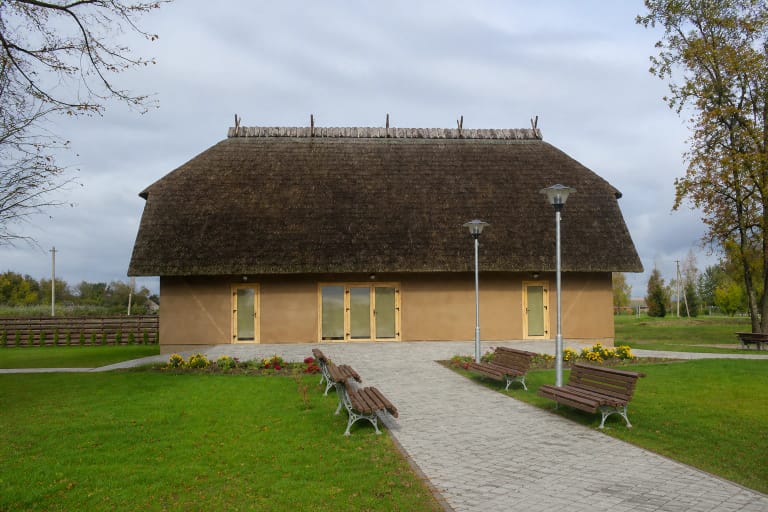 The long low straw-roofed Upytė Traditional Crafts Centre with clay-coloured walls, a straight path, benches, and lamps