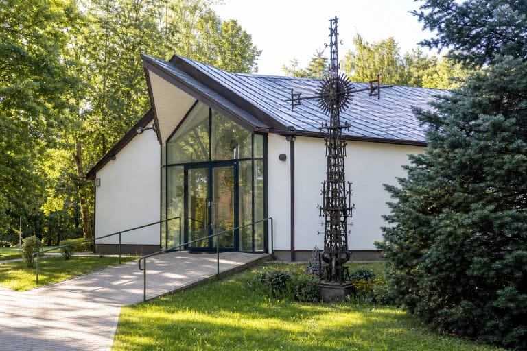 Balta V. K. Jonyno galerija su trikampiu stikliniu įėjimu, nuožulniu metaliniu stogu, pandusu ir aukšta metalo kryžiaus skulptūra žaliame sode
