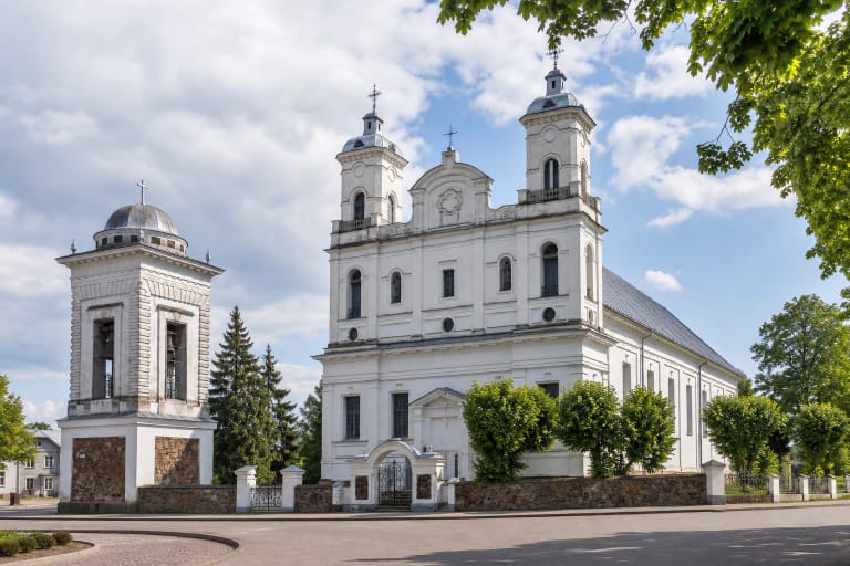 White Vabalninkas Church of the Assumption with two compact facade towers and a detached domed belfry beside its fieldstone churchyard wall