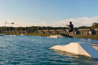 Vandenlentininkas 313 Cable Park trasoje šoka nuo baltos figūros, virš žalio vandens matyti lynų sistema ir mediniai nameliai