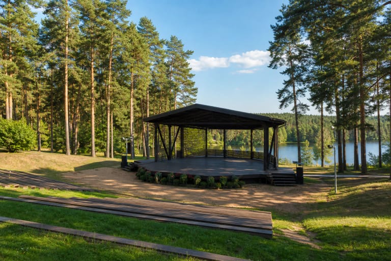 Varėnos dainų slėnio vasaros scena tarp pušų ir prie vandens esančios žiūrovų erdvės