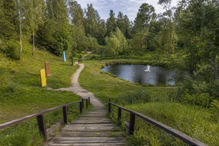 Timber steps descend into green Veiviržėnai Valley, with a fountain pond, walking path and colourful door installations below
