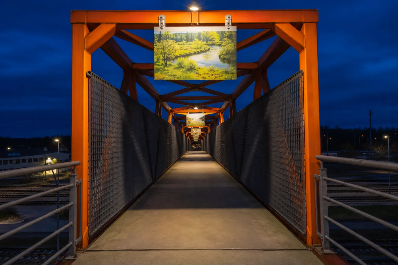 Illuminated orange structure of Viaduko Gallery with exhibition panels suspended above the walkway in Kazlų Rūda