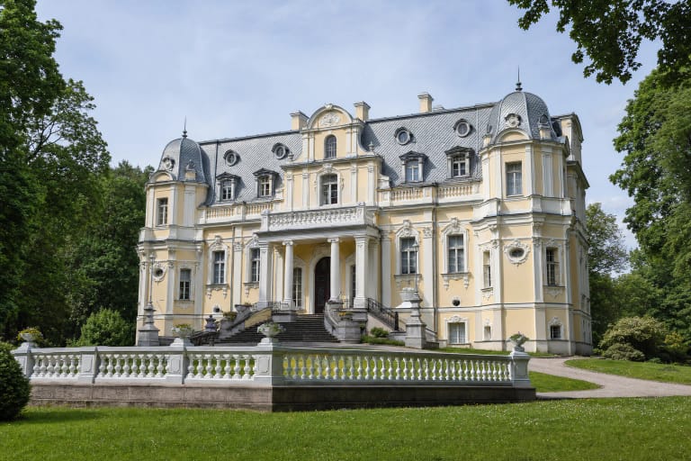 Puošnus gelsvas Vileišių rūmų neobaroko fasadas su laiptais, baliustrada ir pilku mansardiniu stogu Antakalnio žalumoje