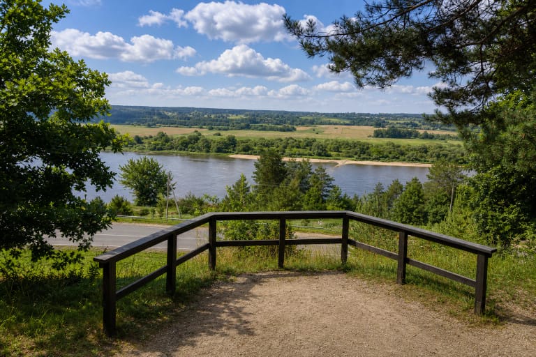 Vilkijos apžvalgos aikštelės žvyro aikštė su žemu mediniu turėklu, keliu apačioje ir Nemuno slėnio panorama