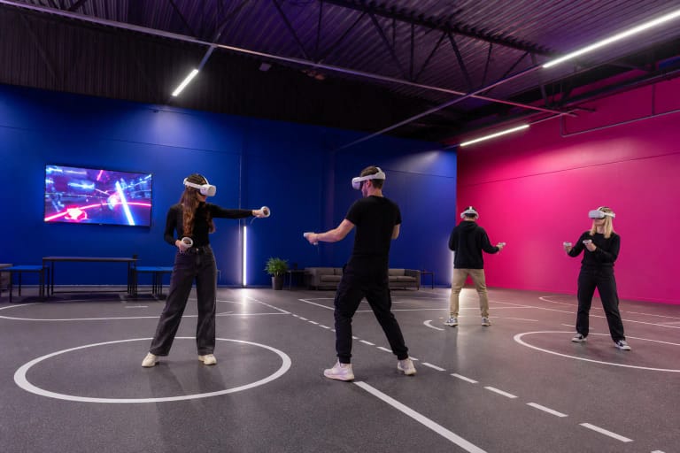 VR ARENA Panevėžys salėje žaidžiantys lankytojai su baltais virtualios realybės akiniais mėlynoje ir rožinėje erdvėje