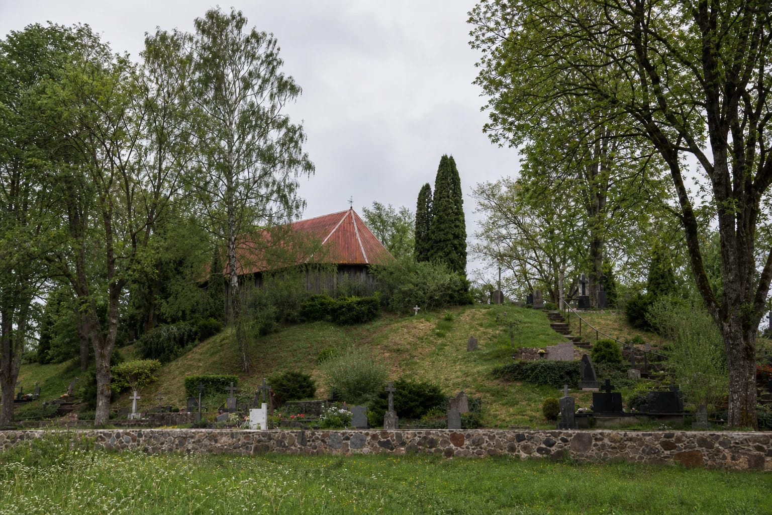 Raktuvė Chapel on Žagarė I Hillfort, surrounded by trees and a historic cemetery above the grassy slope