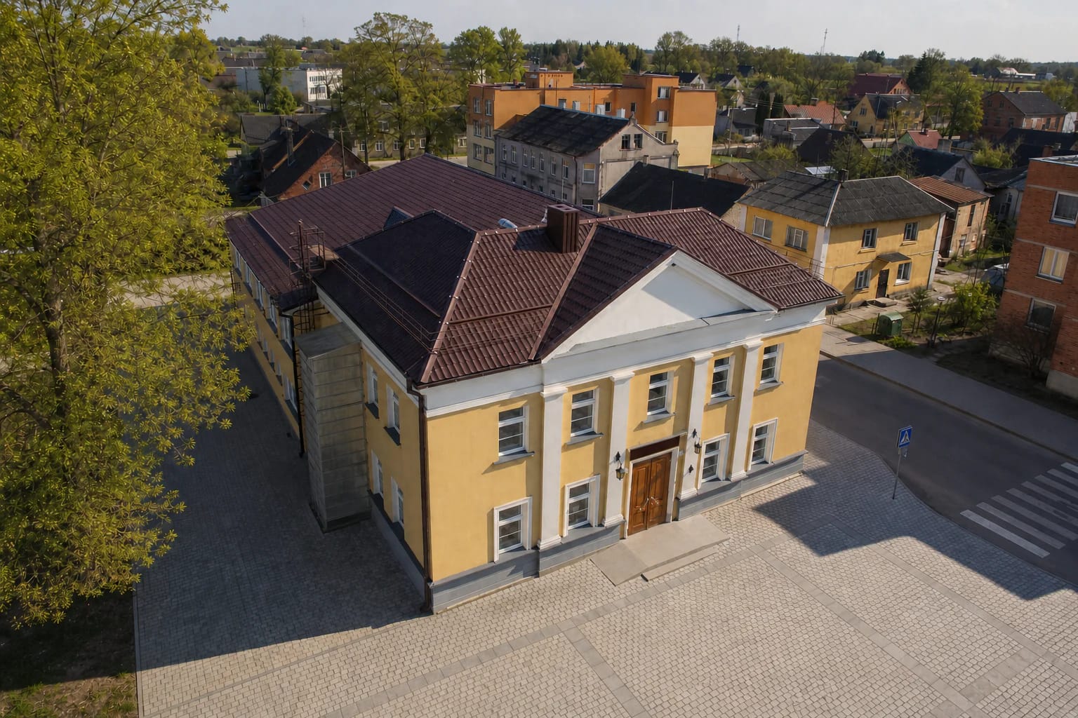 Pale yellow Žagarė Cultural Centre with white trim, red roof, and a paved forecourt
