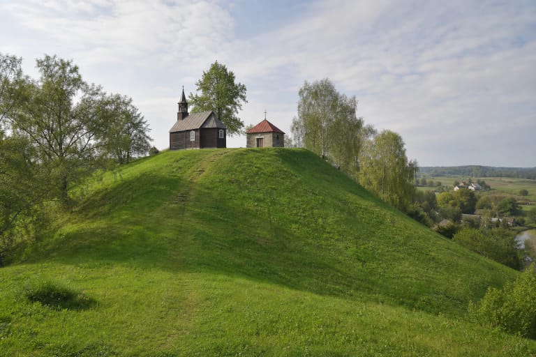 Žalias Žemaičių Kalvarijos piliakalnio šlaitas ir viršuje stovinčios medinė bei mūrinė Kryžiaus kelio koplyčios