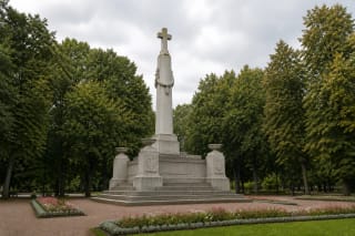 Šviesus devynių metrų paminklas „Žuvome dėl Tėvynės“ su kryžiumi, laiptuotu postamentu ir urnomis Ramybės parko gėlyne